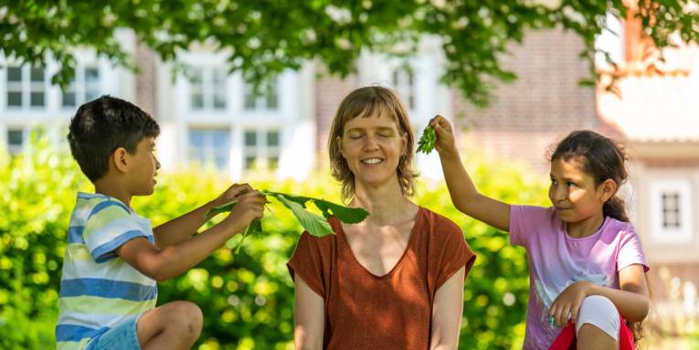 Eine Personen sitzt im Grünen und lauscht den Geräuschen von Blättern, mit denen zwei Kinder ihr zufächern